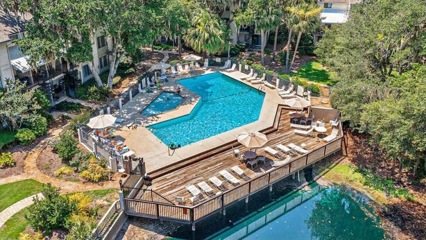 A large outdoor swimming pool surrounded by lounge chairs and umbrellas, with a wooden deck and a small bridge connecting to another pool area. - Home away from Home. Fresh, Bright Condo just steps away from the beach (Hilton Head Island)