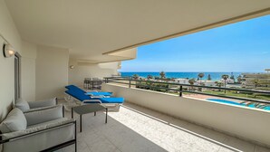 Ein Balkon mit blauen Lounge-Stühlen, einem kleinen Tisch und Blick auf das Meer und Gebäude.