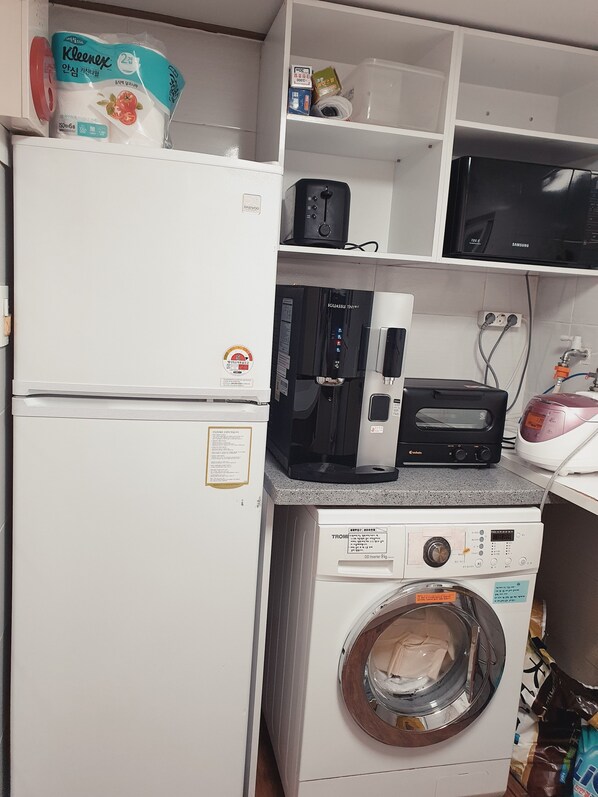 A compact kitchen with a white refrigerator, a black coffee maker, a microwave, a toaster, and a washing machine. - IRich House (Seoul)