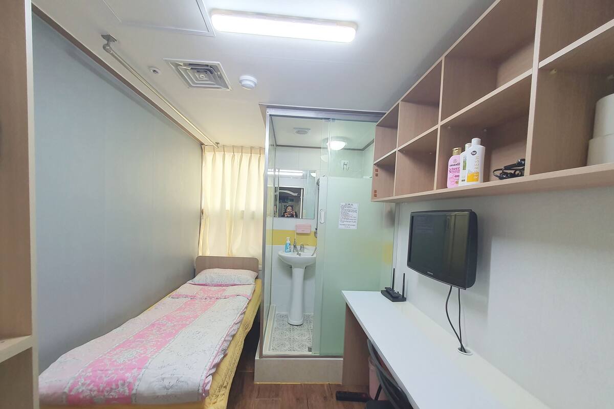 A compact room with a bed, a desk, a television, and a shelf with toiletries.