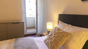A neatly made bed with a pillow and a blanket, a bedside lamp, and a door leading to an outdoor area. - Apartment Blaue Adria (Split)