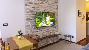 A living room with a stone wall, a flat-screen TV mounted on it, a dining table with yellow tablecloth, and a small side table. - Apartment Blaue Adria (Split)