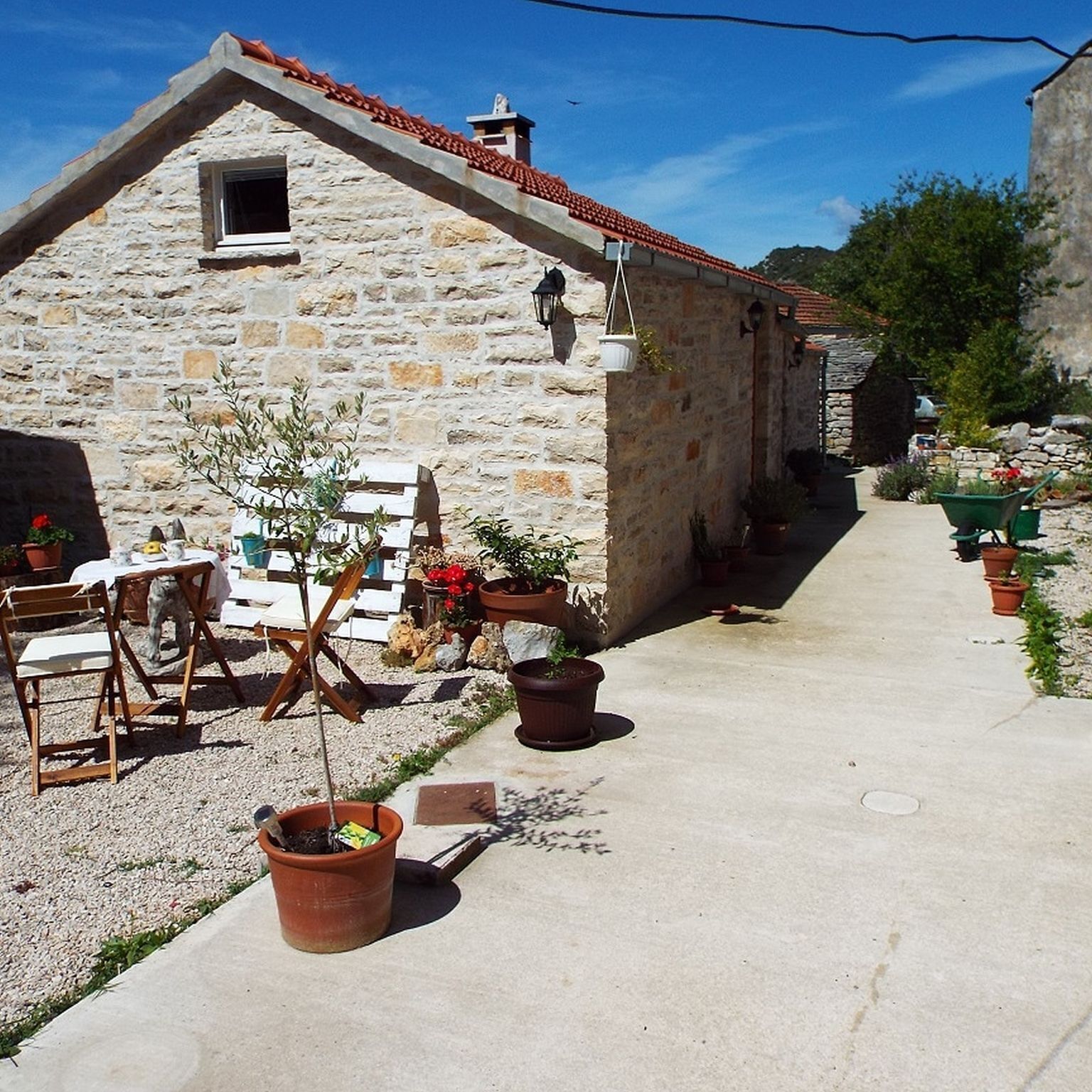 A stone building with a sloped roof, a small window, and a garden area with potted plants and outdoor furniture.