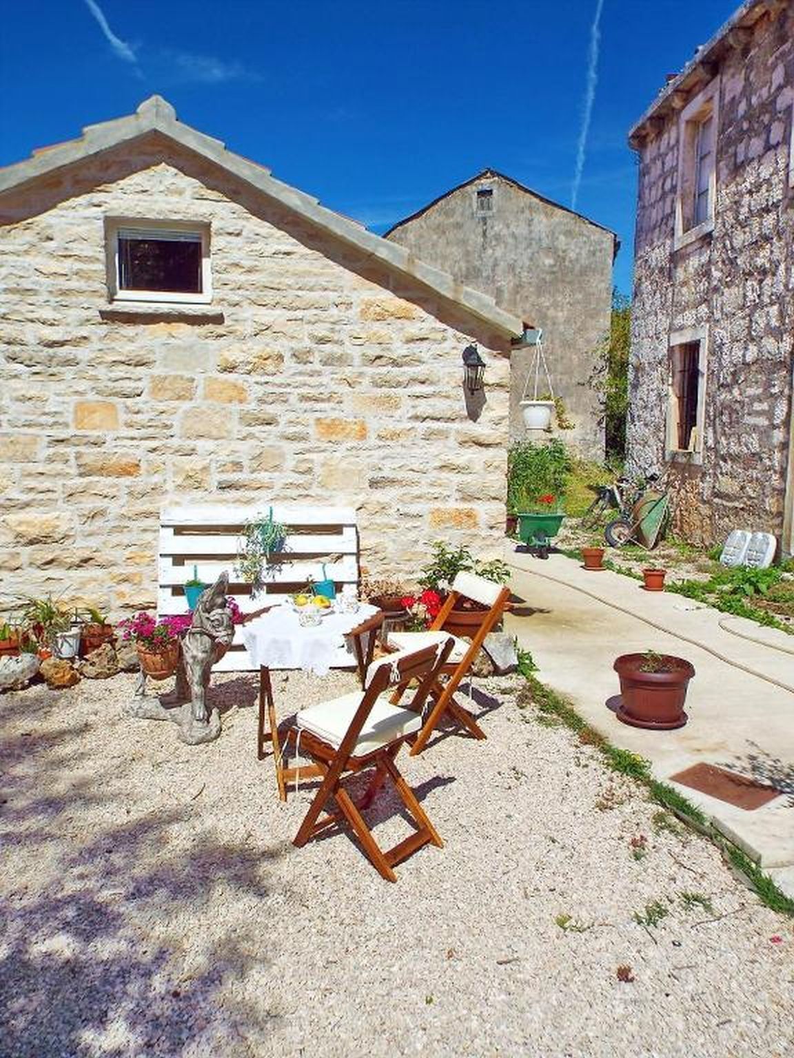 A stone building with a small balcony, outdoor seating with wooden chairs and a table, and potted plants.