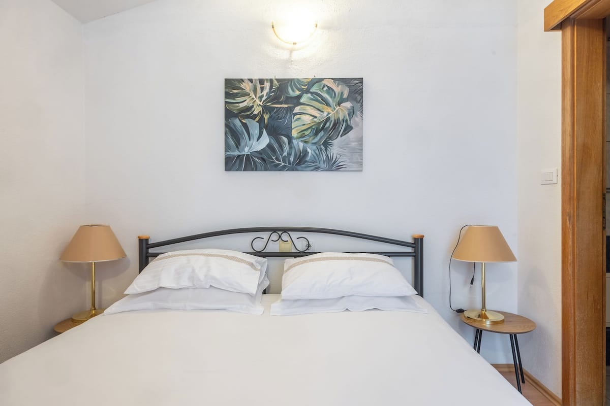 Una cama bien tendida con sábanas blancas, dos lámparas de noche y una obra de arte con hojas tropicales en la pared.
