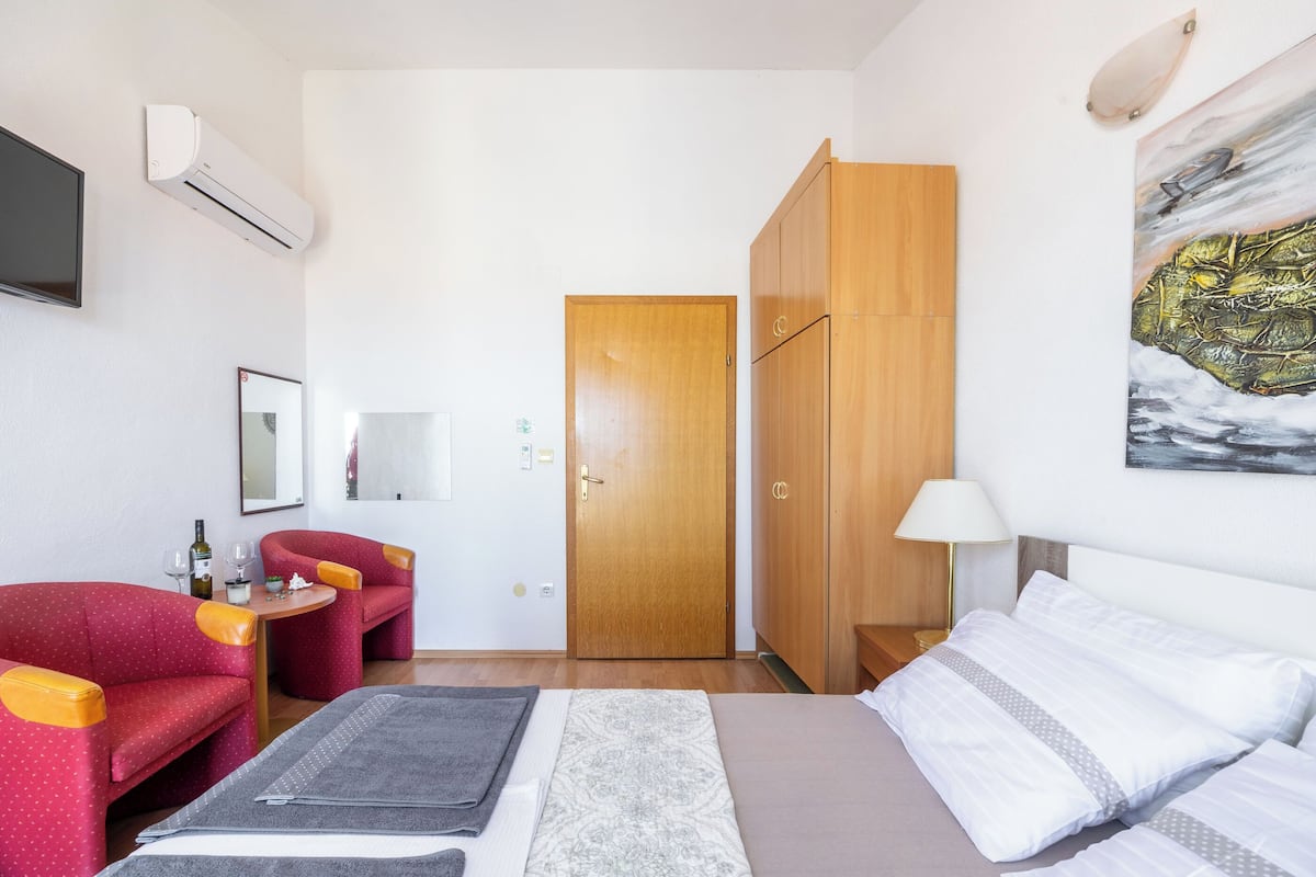 Una habitación de hotel con cama, un sofá rojo, un armario de madera, un cuadro en la pared y un televisor montado en la pared.
