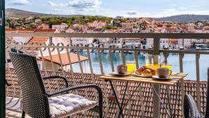 A balcony with wicker chairs, a table with breakfast items, and a view of a marina and town. - Apartment Magical View (Milna)