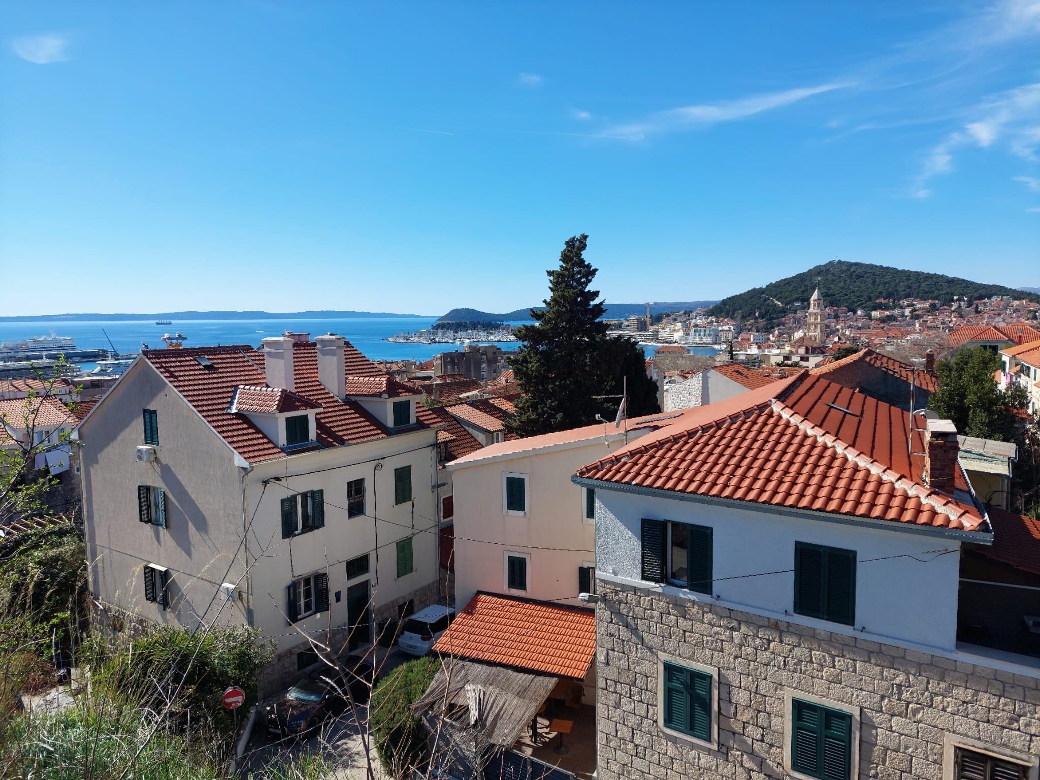 A coastal town with terracotta-roofed buildings, a clear blue sky, and a view of the sea.