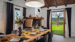 Ein Essbereich mit einem langen Tisch, gedeckt für eine Mahlzeit, einem Kamin und Blick auf eine grüne Landschaft durch eine offene Tür.