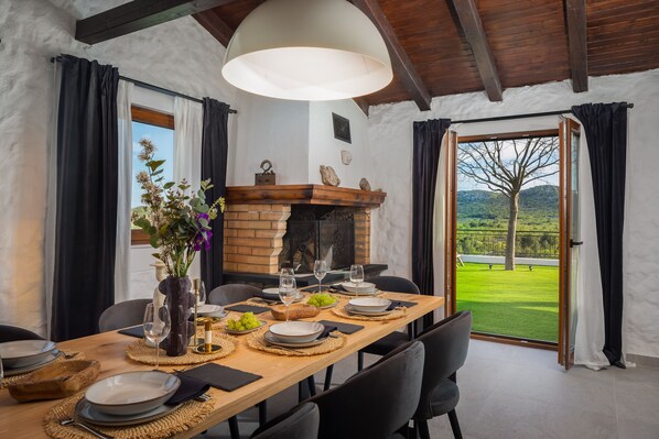 Een eethoek met een lange tafel gedekt voor een maaltijd, een open haard en een uitzicht op een groen landschap door een open deur.
