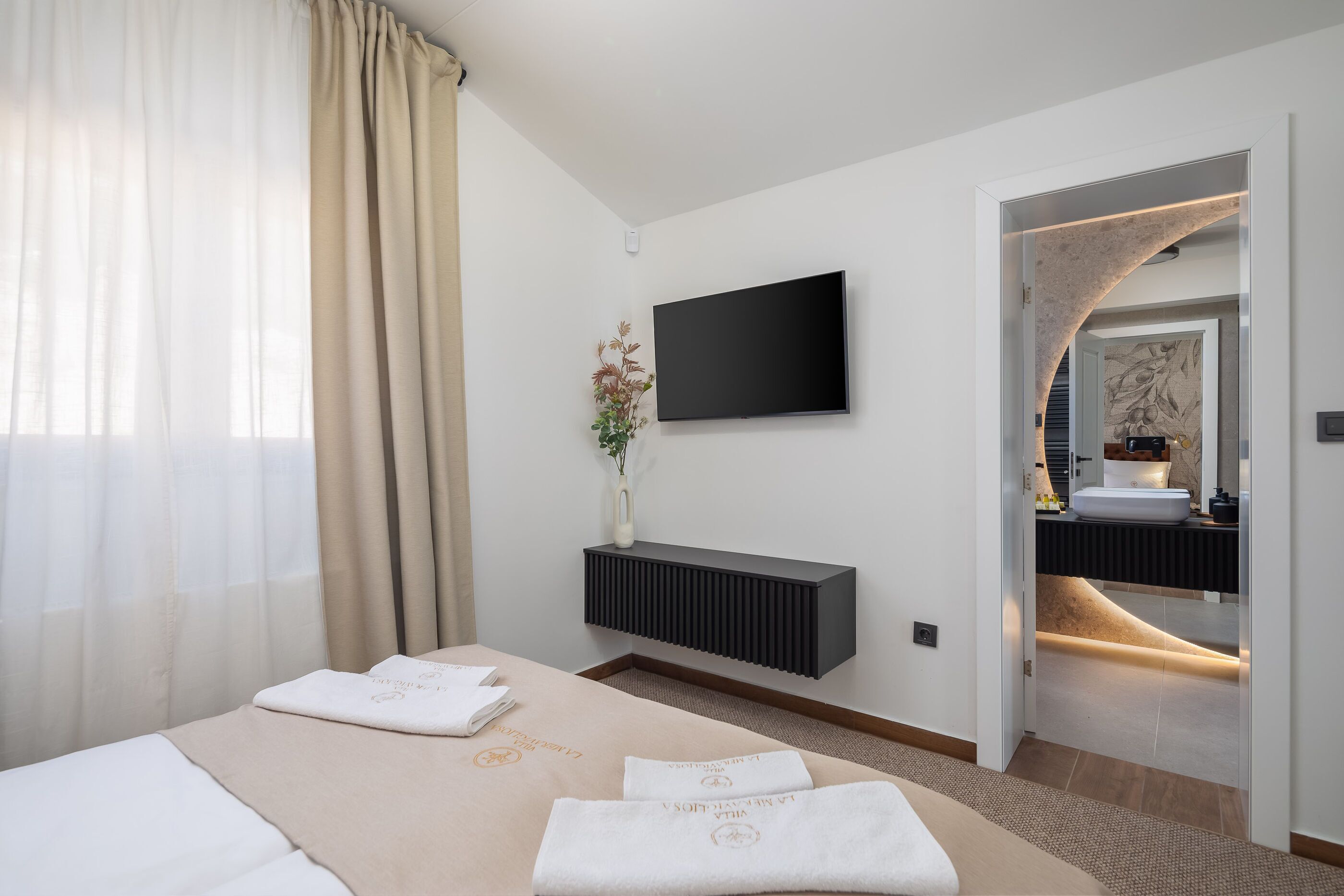 A modern hotel room with a large bed, a flat-screen TV mounted on the wall, a bedside table with a vase of flowers, and a bathroom visible through an open door.