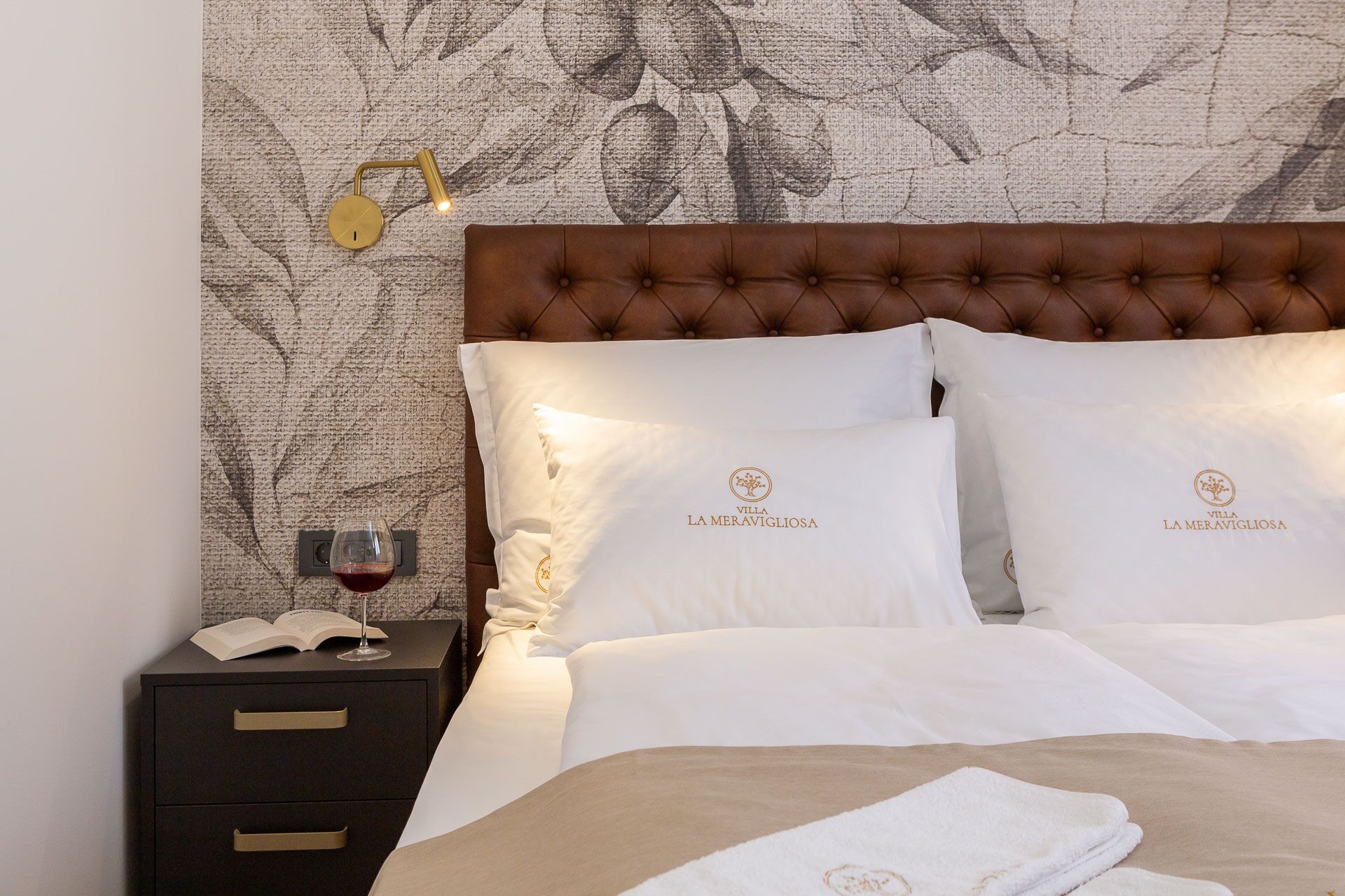 Ein Hotelzimmer mit einer gepolsterten Kopfteil, weißer Bettwäsche und einem Nachttisch mit einem Weinglas und einem Buch.