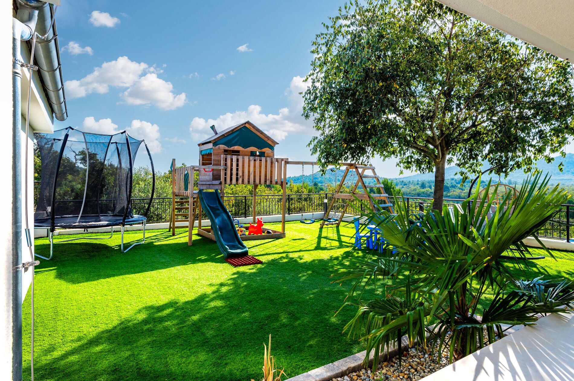 A backyard with a trampoline, a wooden playhouse, and a swing set.