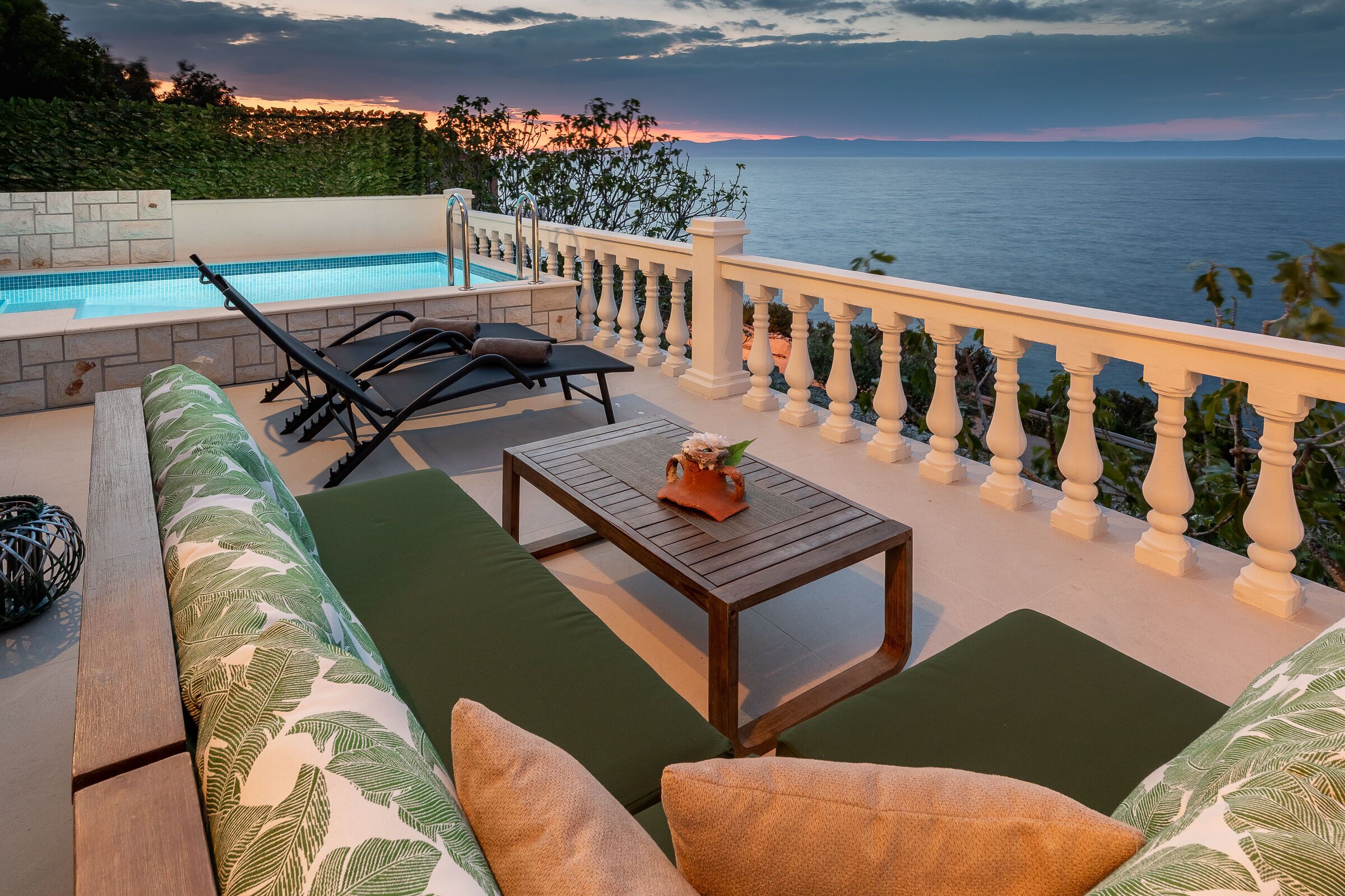 Uma área na piscina com vista para o mar, uma mesa de madeira e um sofá com almofadas verdes.