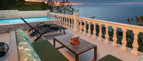 Uma área na piscina com vista para o mar, uma mesa de madeira e um sofá com almofadas verdes.