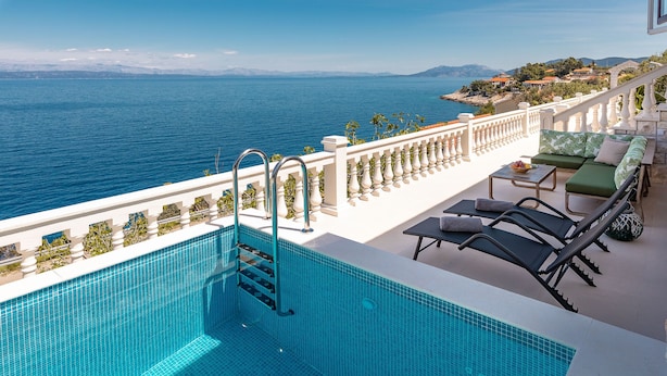 Una piscina con vistas al mar, una barandilla blanca y un área de estar con mesa y sillas.