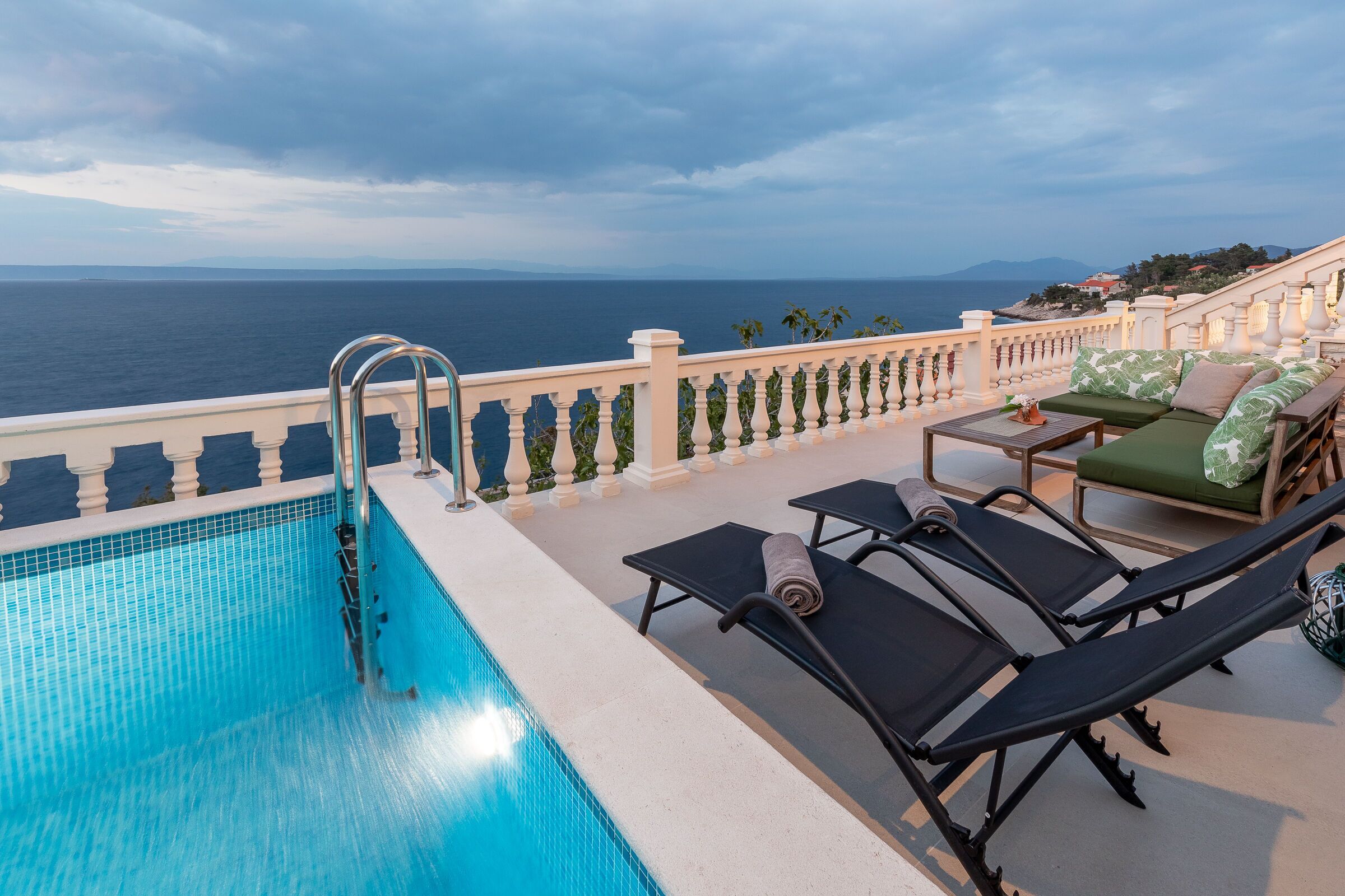 A poolside area with a view of the sea, lounge chairs, and a staircase leading to an upper level.