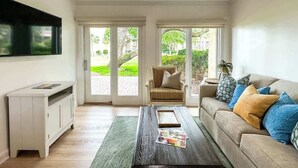 Una sala de estar con sofá, mesa de centro, televisor y puerta corrediza de vidrio que da a un jardín.