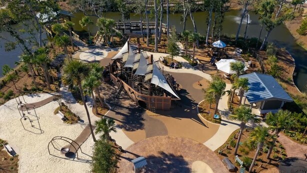 Vista aérea de un parque con zona arenosa, un elemento acuático y un pabellón sombreado.