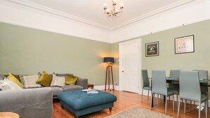 A living room with a grey sofa, blue ottoman, and a dining area with a table and chairs. - Spinners House (Dartmouth)
