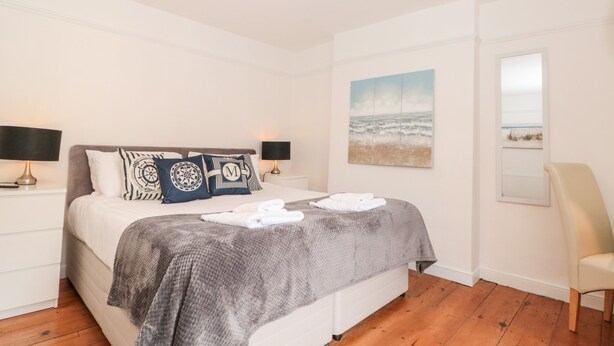 A bedroom with a bed, two bedside lamps, a painting of the ocean, a chair, and a wooden floor. - Spinners House (Dartmouth)