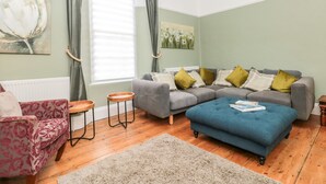 A living room with a grey sofa, blue ottoman, and a pink armchair. There are two small wooden tables and a large painting on the wall. - Spinners House (Dartmouth)
