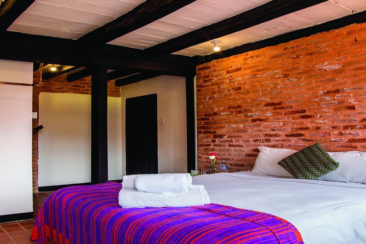 Quarto com parede de tijolos, cama com lençóis brancos e manta listrada colorida.
