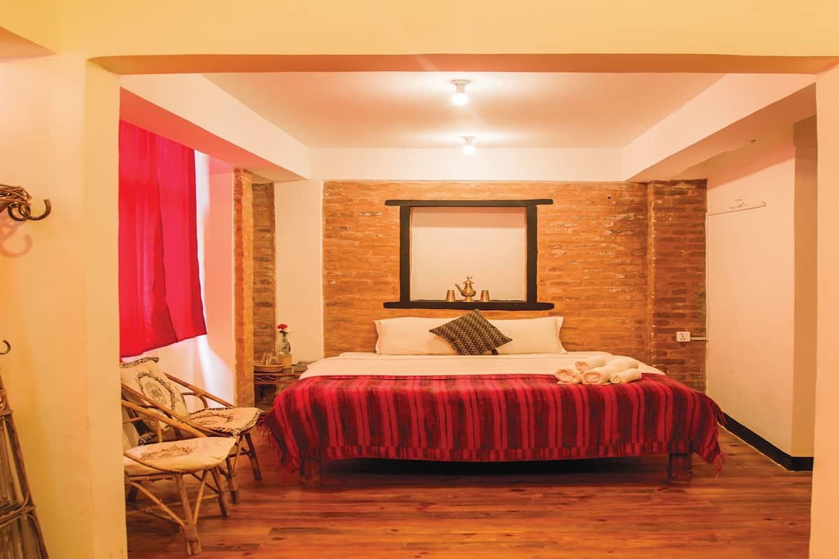 Quarto com piso de madeira, cama com cobertor vermelho, espelho na parede e uma cadeira.