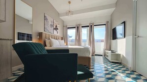 Ein Hotelzimmer mit Bett, Sessel, Fernseher und einem Balkon mit Vorhängen sowie einem gemusterten Boden.