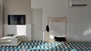 Ein Zimmer mit an der Wand befestigtem Fernseher, einer Tür, einer wandmontierten Klimaanlage, einem Wäscheständer mit Kleidung und einem gemusterten Boden.