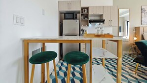 Eine kompakte Küche mit Mikrowelle, Kühlschrank und Holzschränken. Zwei grüne Barhocker stehen an einem Tisch mit Marmorplatten. Der Boden ist mit einem geometrischen Fliesenmuster versehen.