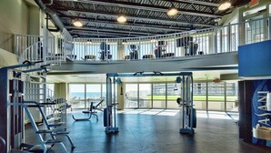 Ein Fitnessraum mit verschiedenen Trainingsgeräten, einem Balkon und großen Fenstern mit Blick ins Freie.