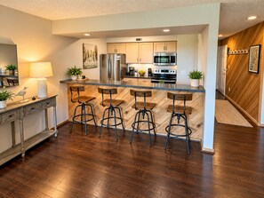 Una cocina moderna con una isla central, taburetes de bar, electrodomésticos de acero inoxidable y suelo de madera.