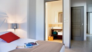 A hotel room with a bed, a nightstand, a lamp, a bathroom with a mirror, and a door leading to another room.