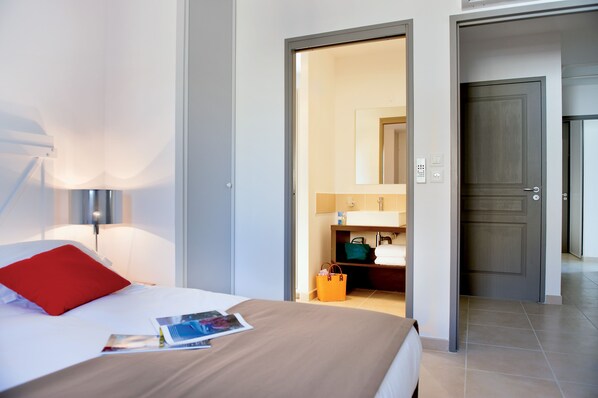 A hotel room with a bed, a nightstand, a lamp, a bathroom with a mirror, and a door leading to another room.