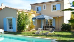 A swimming pool with a sun umbrella, a dining table with chairs, and a house with blue shutters.