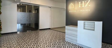 Egy modern szállodai előcsarnok, geometrikus mintázatú padlóval, recepciós pulttal és a falra szerelt „Hotel Z-TRIHOA” felirattal.