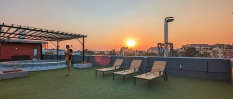 Terraço no telhado com vista para o pôr do sol, cadeiras de descanso e uma estrutura de pérgula.