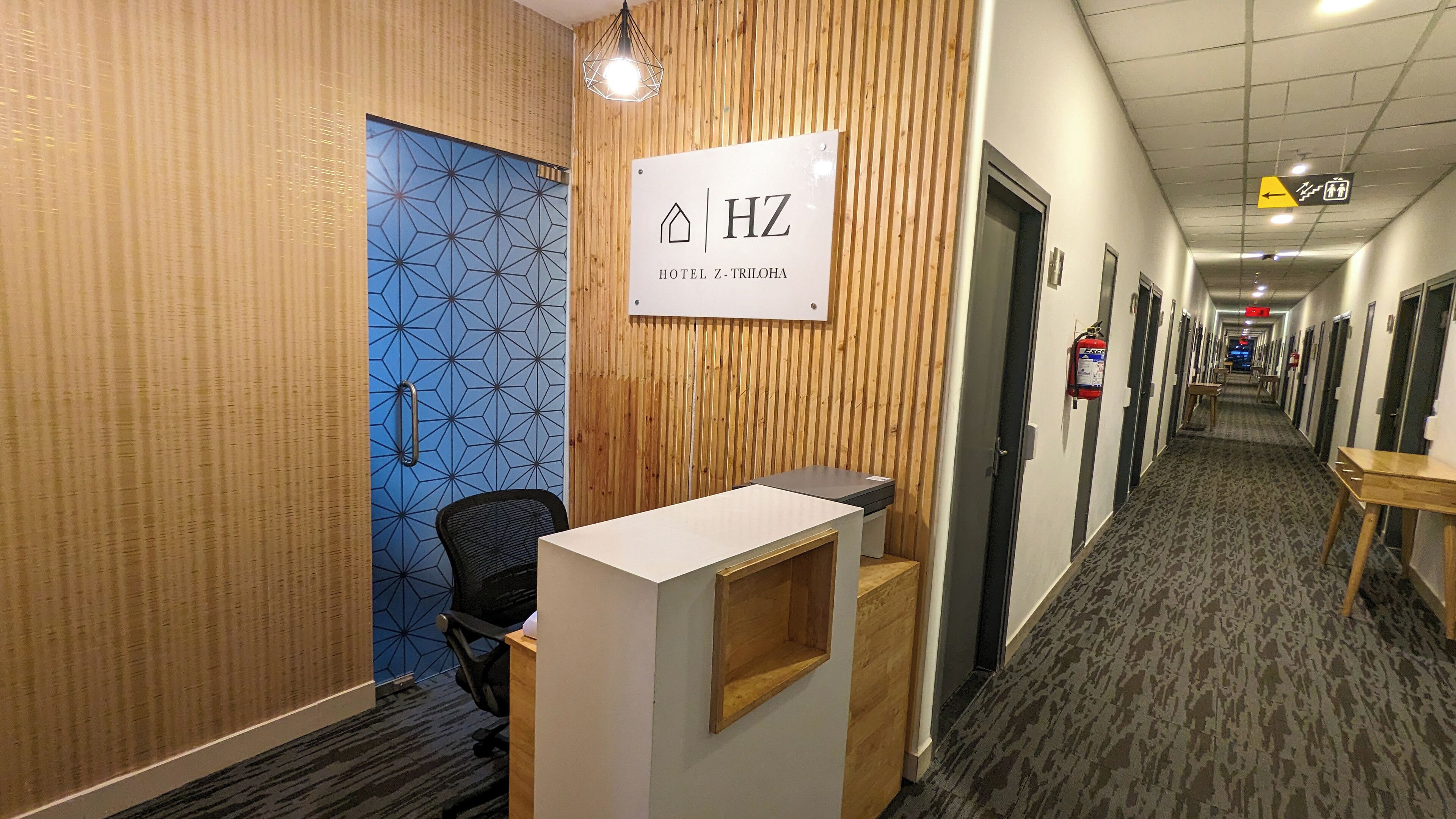 Zona de recepción de hotel con un cartel que dice HOTEL Z-TRILOA, un mostrador de recepción, una silla y un extintor en la pared.