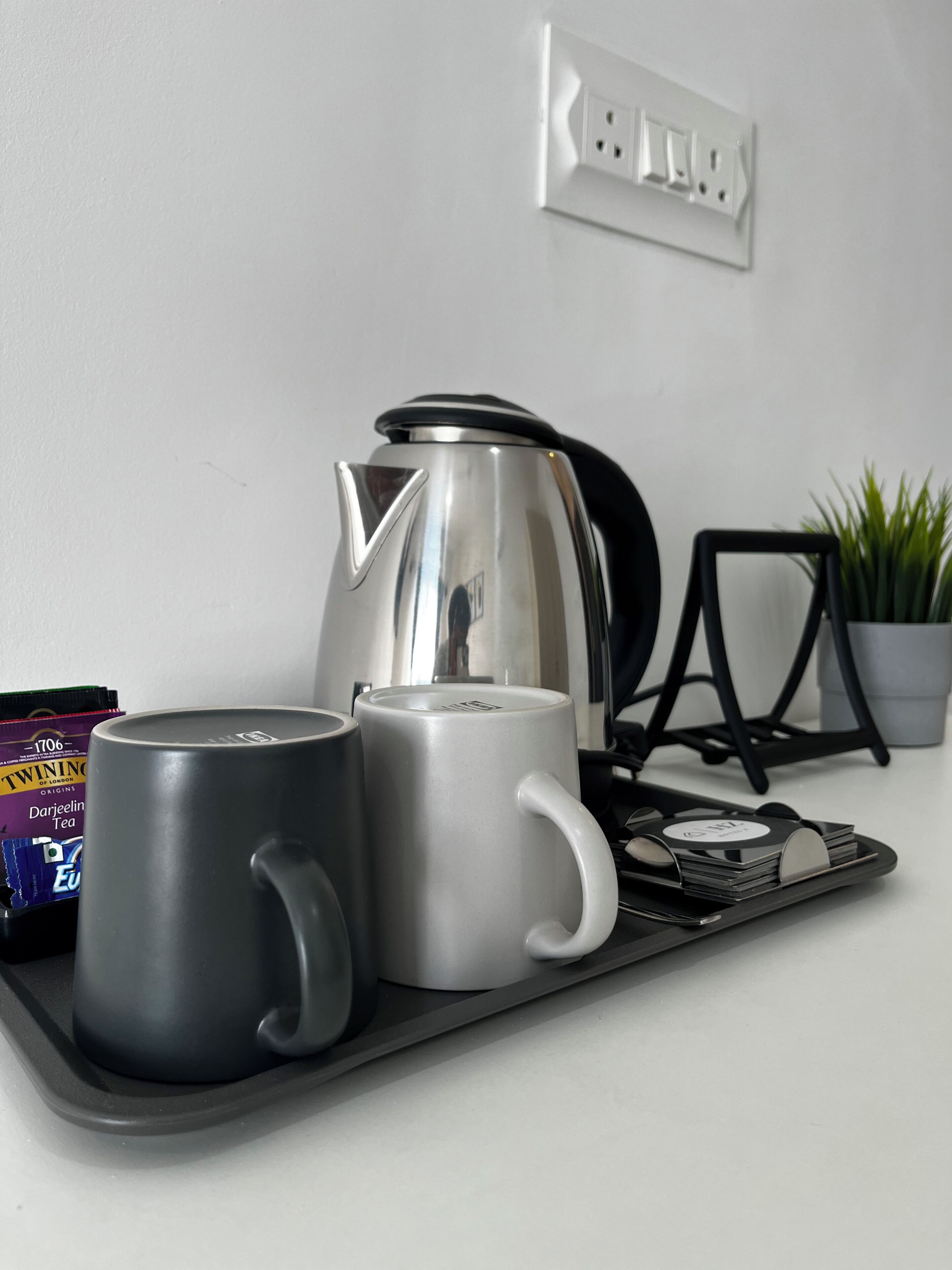 A kitchen counter with a kettle, two mugs, a tray, a plant, and a power outlet.