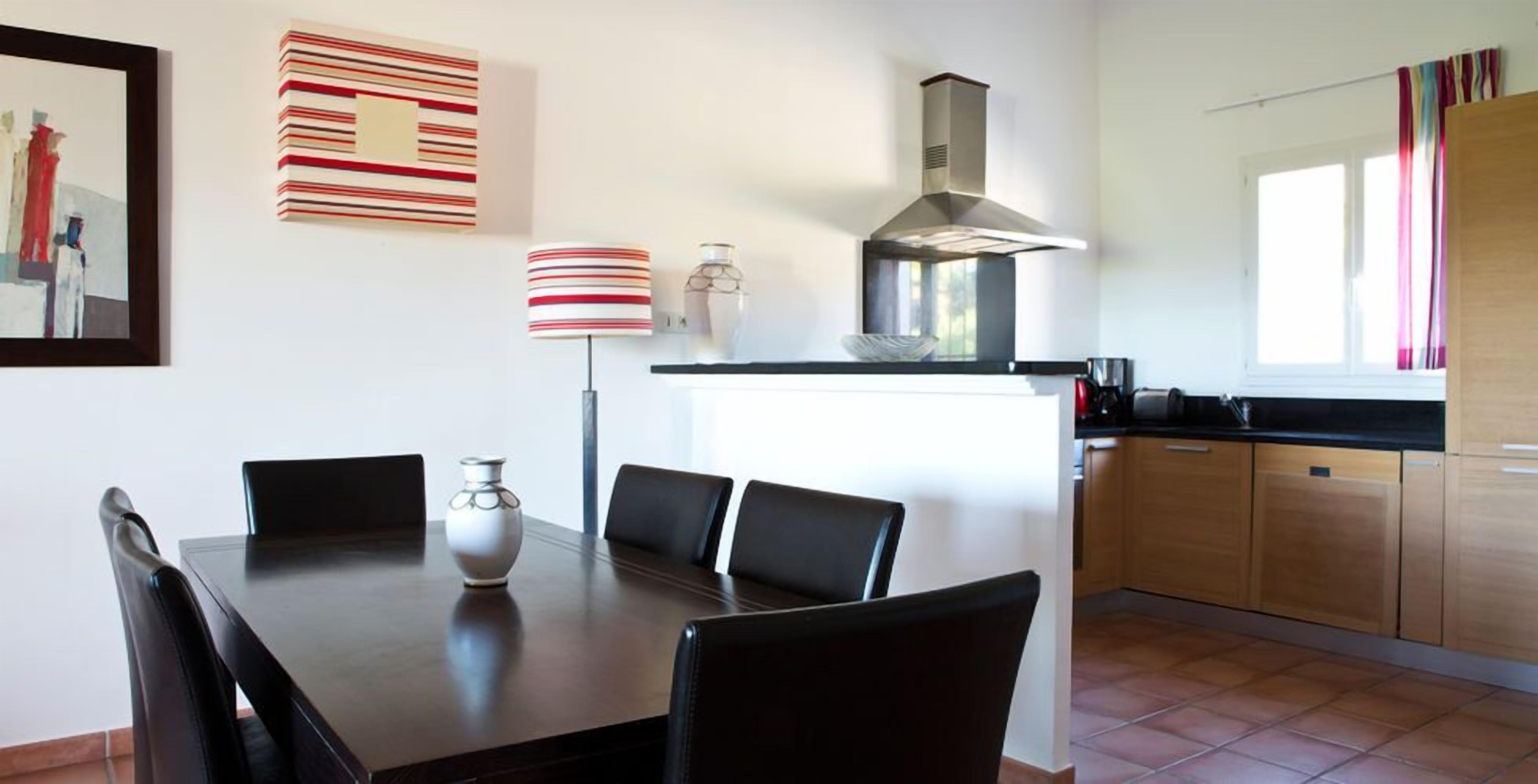 A modern kitchen with a dining area, featuring a central island, black chairs, a vase, and a striped lamp.