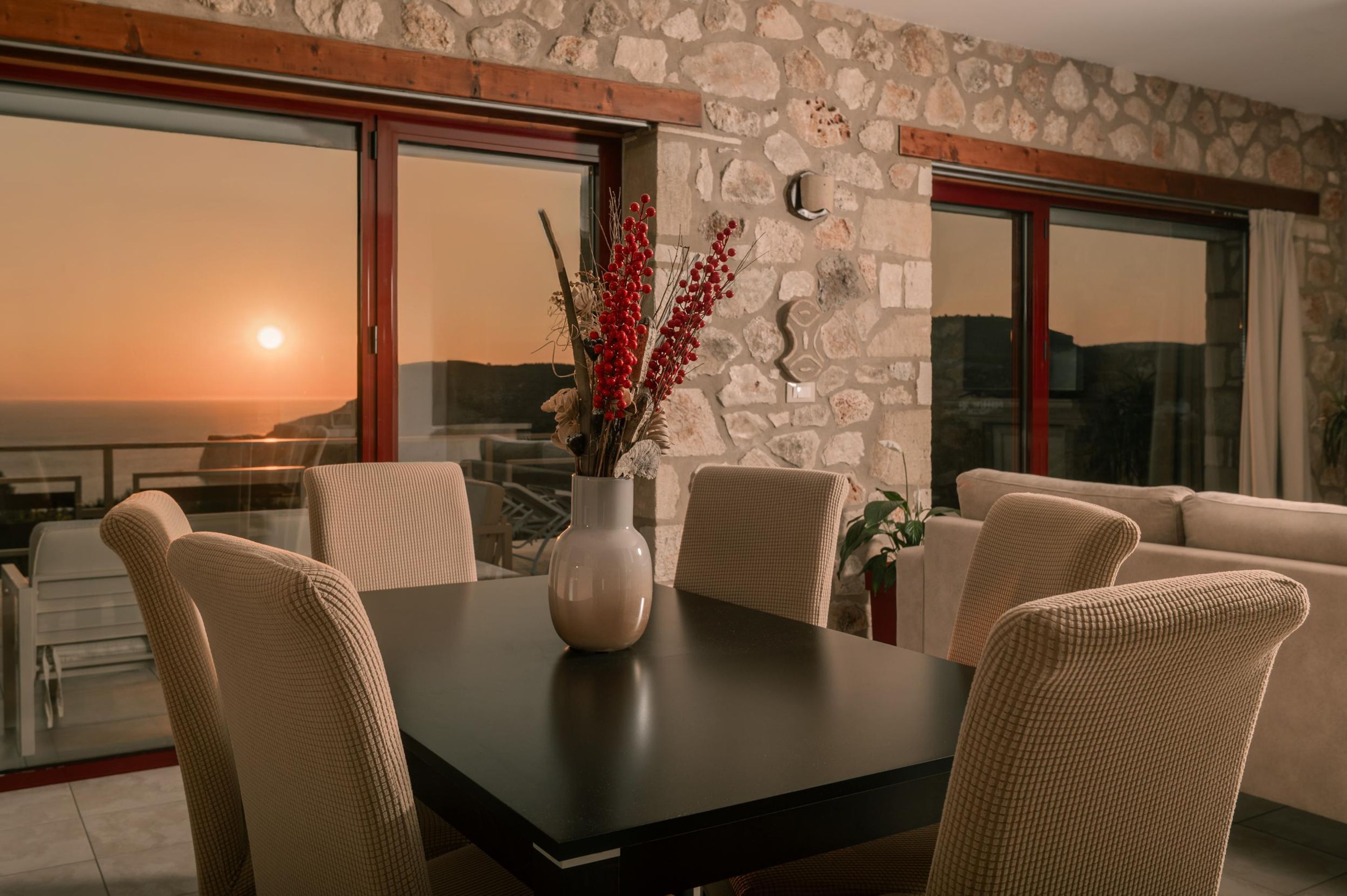 A dining area with a stone wall, a large window with a view of the sea, and a vase with red berries on the table.