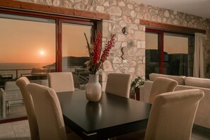 A dining area with a stone wall, a large window with a view of the sea, and a vase with red berries on the table. - Arca Villa - Enchanting Sunset! (Zakynthos)
