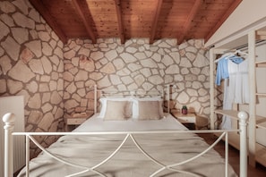 A bedroom with a stone wall, a wooden ceiling, a bed with a metal frame, a nightstand, and a hanging rack with clothes. - Arca Villa - Enchanting Sunset! (Zakynthos)