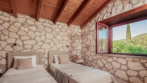 A bedroom with two beds, stone walls, wooden beams, and a window with a view of greenery. - Arca Villa - Enchanting Sunset! (Zakynthos)