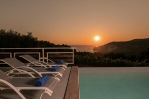 A poolside area with lounge chairs and a view of the sunset over the sea.