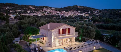 Una villa de piedra con piscina, zona de estar al aire libre y vistas a un pueblo en la ladera.