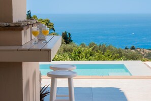 A poolside area with a view of the sea, a table with glasses of orange juice, and a white stool. - Arca Villa - Enchanting Sunset! (Zakynthos)