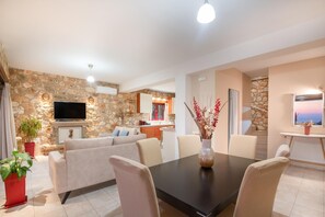 A modern living room with a stone wall, a flat-screen TV, a sofa, and a dining table with chairs. - Arca Villa - Enchanting Sunset! (Zakynthos)