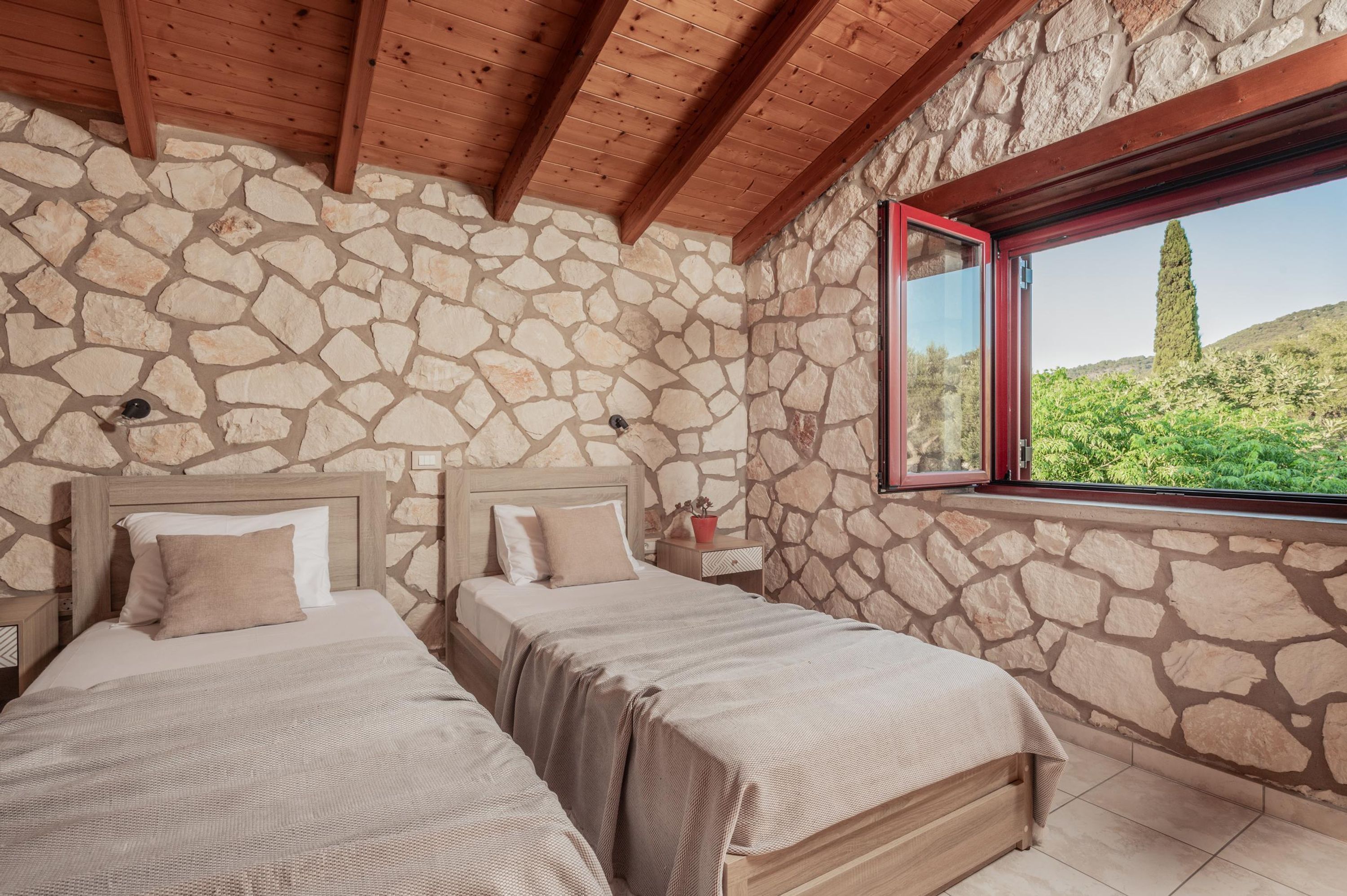A bedroom with two beds, stone walls, wooden beams, and a window with a view of greenery.